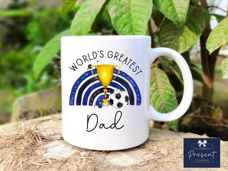Puede incluir: Taza de cer&aacute;mica blanca con un dise&ntilde;o de arco&iacute;ris azul y blanco, un trofeo dorado y un bal&oacute;n de f&uacute;tbol. El texto "World's Greatest Dad" est&aacute; impreso en la taza.