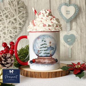 May include: White ceramic mug with a red handle, featuring a snow globe design with a ballerina inside. The snow globe depicts a Christmas tree and a nutcracker. The mug is topped with whipped cream and candy canes, and the name "Thomas" is written on the front.