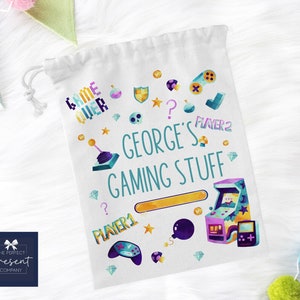 Op de afbeelding: Een witte trekkoordtas met een kleurrijk gamingthema. De tas heeft een blauwe en witte tekst die "Game Over" en "George's Gaming Stuff" zegt. De tas is versierd met een kleurrijke gamecontroller, joystick en andere gamingpictogrammen.