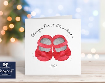 May include: A white Christmas card with red baby shoes and the text "Ebony's First Christmas 2022".
