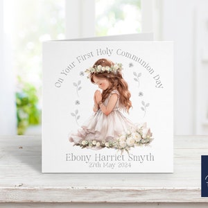 May include: A white card with a watercolor illustration of a girl in a white dress kneeling with her hands clasped in prayer. The card has the text "On Your First Holy Communion Day" and "Ebony Harriet Smyth 27th May 2024".