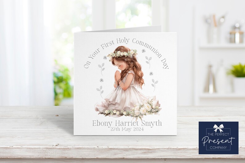 May include: A white card with a watercolor illustration of a girl in a white dress kneeling with her hands clasped in prayer. The card has the text "On Your First Holy Communion Day" and "Ebony Harriet Smyth 27th May 2024".