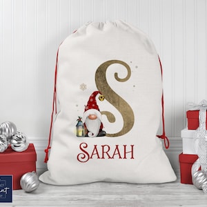 May include: White Christmas gift bag with a red drawstring. The bag features a gold glitter letter "S" and a gnome with a red hat and beard. The name "SARAH" is written in red cursive below the gnome.