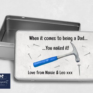 Puede incluir: Una lata de metal plateada con fondo blanco y el texto "When it comes to being a Dad... You nailed it!" Se muestra un martillo azul y plateado y clavos plateados. El texto "Love from Maisie & Leo xxx" está en la parte inferior de la lata.