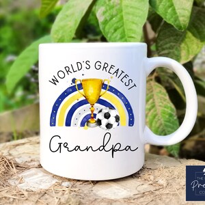 Puede incluir: Taza de cer&aacute;mica blanca con un dise&ntilde;o de arco&iacute;ris azul y amarillo, un trofeo dorado y un bal&oacute;n de f&uacute;tbol. El texto "World's Greatest Grandpa" est&aacute; impreso en la taza.