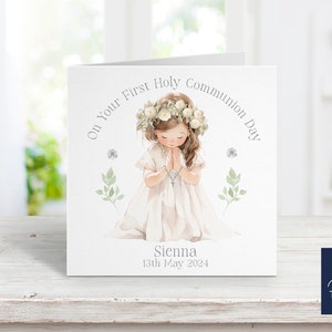 May include: A white card with a watercolor illustration of a girl in a white dress with a flower crown, praying. The text on the card reads "On Your First Holy Communion Day" and "Sienna 13th May 2024".