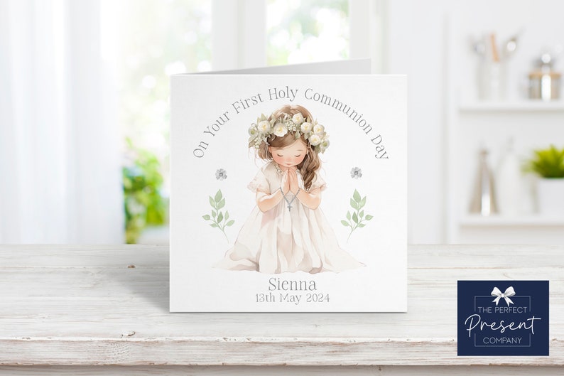 May include: A white card with a watercolor illustration of a girl in a white dress with a flower crown, praying. The text on the card reads "On Your First Holy Communion Day" and "Sienna 13th May 2024".