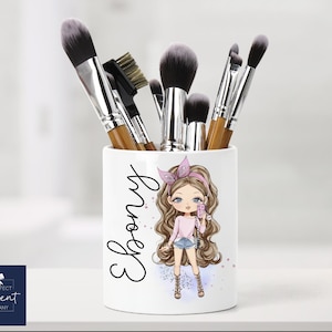 May include: White ceramic makeup brush holder featuring a cartoon girl design. The girl is wearing a pink shirt, blue jeans, and a pink headband. The holder has the name "Ebony" written on it in black.