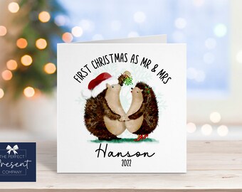 May include: A white card with a Christmas-themed illustration of two hedgehogs under a sprig of mistletoe. The hedgehog wearing a Santa hat is holding the other hedgehog's hand. The text on the card reads "First Christmas as Mr & Mrs Hanson 2022".