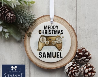 May include: A white round wooden ornament with a white circle in the center. The circle has a watercolor-style image of a gold video game controller with the text "Merry Christmas" above it and the name "Samuel" below it.