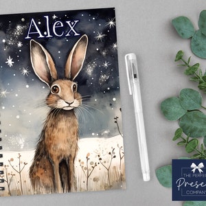 May include: A spiral bound notebook with a watercolor illustration of a brown hare in a snowy field under a starry night sky. The name "Alex" is written in blue at the top of the notebook.