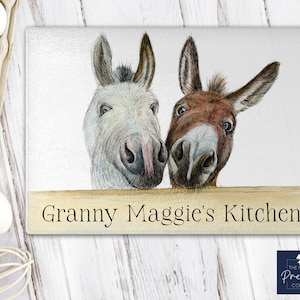 Puede incluir: Un burro blanco y marrón asomándose por encima de un letrero de madera que dice "Granny Maggie's Kitchen".