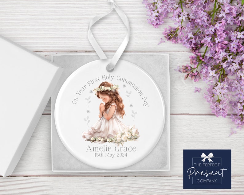 May include: White ceramic ornament with a girl in a white dress praying. The ornament has the text "On Your First Holy Communion Day" and "Amelie Grace 15th May 2024".