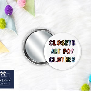 May include: A round compact mirror with a white lid that says "CLOSETS ARE FOR CLOTHES" in colorful letters. The mirror is open and the lid is laying next to it.