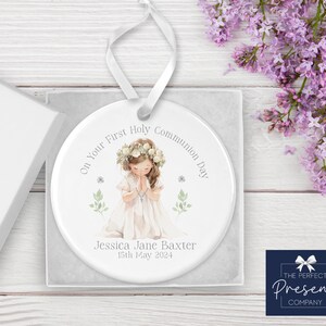 May include: White ceramic ornament with a watercolor illustration of a girl in a white dress praying. The ornament has the text "On Your First Holy Communion Day" and "Jessica Jane Baxter 15th May 2024".