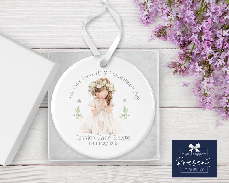 May include: White ceramic ornament with a watercolor illustration of a girl in a white dress praying. The ornament has the text "On Your First Holy Communion Day" and "Jessica Jane Baxter 15th May 2024".