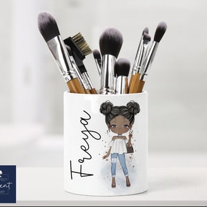 May include: White ceramic makeup brush holder with a cartoon illustration of a Black girl with brown hair in pigtails. The holder has the name "T'reya" written on it in black.