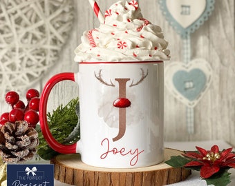 May include: White ceramic mug with a red rim and handle. The mug features a watercolor reindeer design with a brown letter "J" and the name "Joey" printed in red. The mug is topped with whipped cream and candy canes.