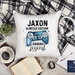 Könnte beinhalten: Weißes quadratisches Kissen mit einem blauen Videospiel-Controller-Design im Aquarellstil. Der Text auf dem Kissen lautet "Jaxon Limited Edition Gaming Legend". Das Kissen ist von einer Decke, Kopfhörern und einer Tasse Kaffee umgeben.