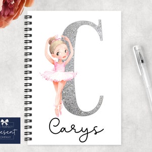 Può includere: Un quaderno a spirale bianco con una lettera C di glitter argentato e un'illustrazione di ballerina. Il nome "Carys" è scritto in corsivo nero sotto la lettera.
