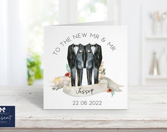 May include: A square wedding card with a watercolor illustration of two grooms in tuxedos. The text reads "TO THE NEW MR & MR" above the figures, with a banner below displaying a name and date. Floral accents and champagne glasses complete the design.
