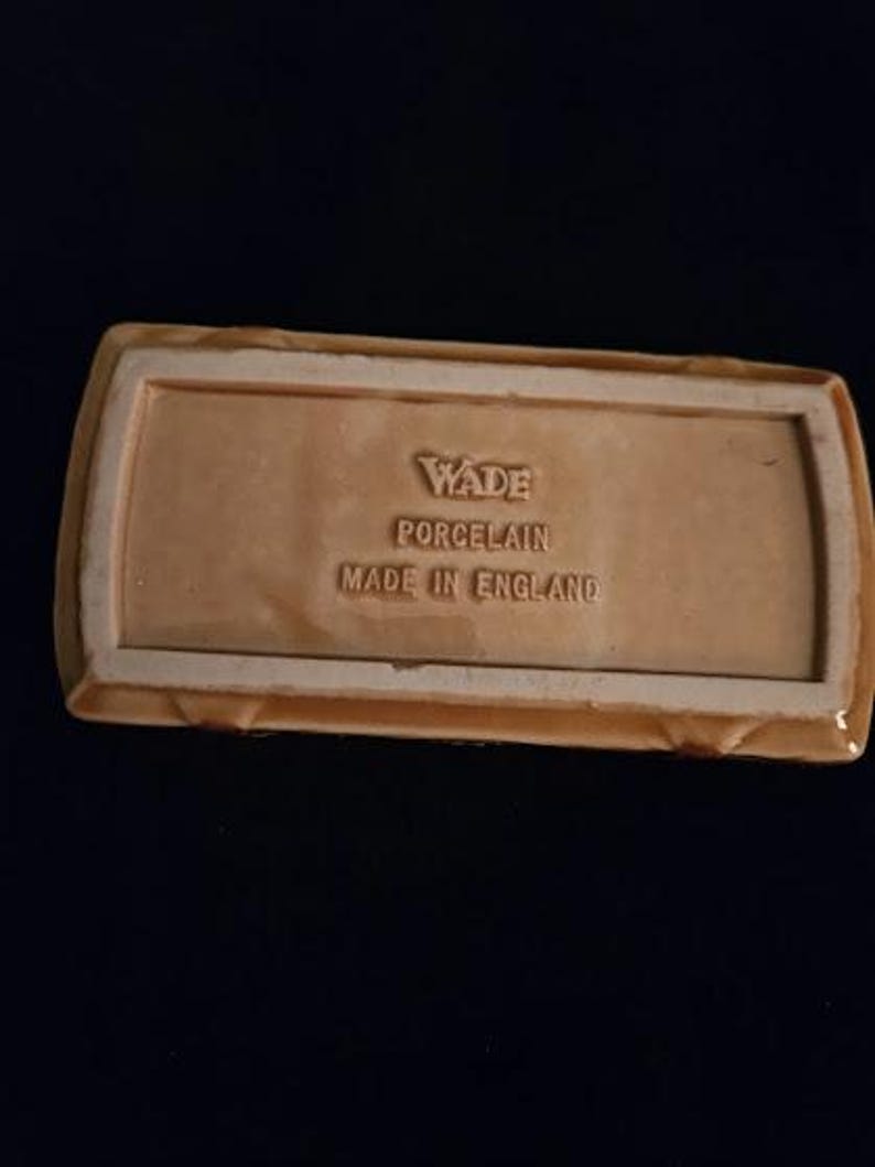 May include: A rectangular, light brown Wade porcelain dish with a raised border. The base features embossed text: "Wade Porcelain Made in England."