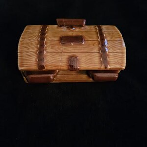 May include: A ceramic treasure chest-shaped container with a wood-grain design. The container is brown with darker brown accents, including straps and a handle. The chest has a rectangular shape with rounded edges.