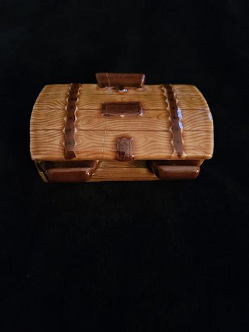 May include: A ceramic treasure chest-shaped container with a wood-grain design. The container is brown with darker brown accents, including straps and a handle. The chest has a rectangular shape with rounded edges.