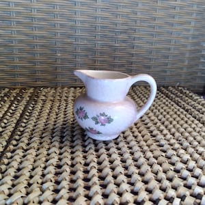 May include: A small, pink ceramic creamer with a handle and spout. The creamer features a floral design with pink roses and green leaves. The creamer is approximately 4 inches tall and has a rounded body.