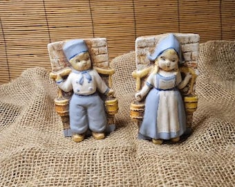 May include: Two ceramic figurines of Dutch children. The boy wears a blue hat, jacket, and pants, holding buckets. The girl wears a blue bonnet and dress, also holding buckets. Both stand in front of brick-like structures.