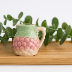 May include: A small, decorative ceramic pitcher with a handle, featuring a green top and a pink, textured body. It sits on a rectangular bamboo tray, with green foliage in the background. The pitcher is likely for decorative purposes.