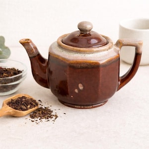 May include: A brown ceramic teapot with a lid, handle, and spout, alongside a small white cup and a glass bowl of tea leaves. A wooden scoop holds more tea leaves. The teapot has a light-colored band around the top and is marked "MADE IN TAIWAN".