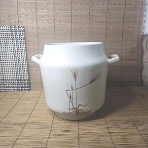 May include: White ceramic container with two handles and a wheat stalk design. The container is cylindrical with a slightly flared base. The wheat design is in shades of brown and tan. The container is on a bamboo mat.