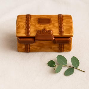 May include: A ceramic treasure chest with a wood-grain texture in shades of brown. The chest has a rectangular shape with a hinged lid, secured by straps and a clasp. A sprig of green leaves rests beside the chest.