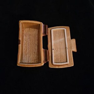 May include: Open, rectangular, ceramic trinket box with a hinged lid. The box and lid are tan with a wood grain pattern. The interior of the box is also tan. The box is on a black background.