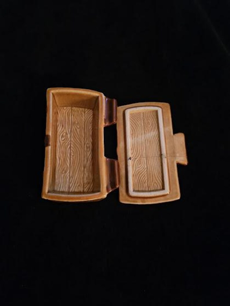 May include: Open, rectangular, ceramic trinket box with a hinged lid. The box and lid are tan with a wood grain pattern. The interior of the box is also tan. The box is on a black background.