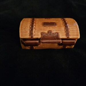 May include: A small, ceramic treasure chest with a wooden plank design. The chest is light brown with darker brown accents, including straps and a clasp. The lid is slightly curved, and the chest sits on a dark background.