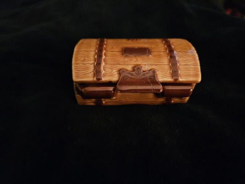 May include: A small, ceramic treasure chest with a wooden plank design. The chest is light brown with darker brown accents, including straps and a clasp. The lid is slightly curved, and the chest sits on a dark background.