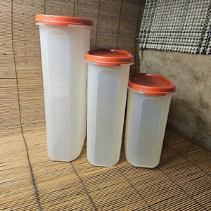May include: Three clear, rectangular food storage containers with orange lids. The containers are different sizes, with the tallest one on the left. The containers are made of a translucent plastic material.