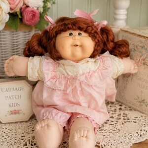 May include: A Cabbage Patch Kids doll with auburn hair in pigtails, wearing a cream-colored floral blouse and a pink ruffled romper. The doll has a soft fabric body, light skin tone, and is positioned next to a sign that reads Cabbage Patch Kids.