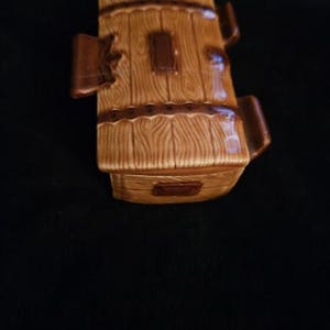 May include: A ceramic barrel-shaped container with a wood-grain design in shades of brown. The container has a rectangular opening and two handles. The design includes dark brown accents and a decorative band around the middle.