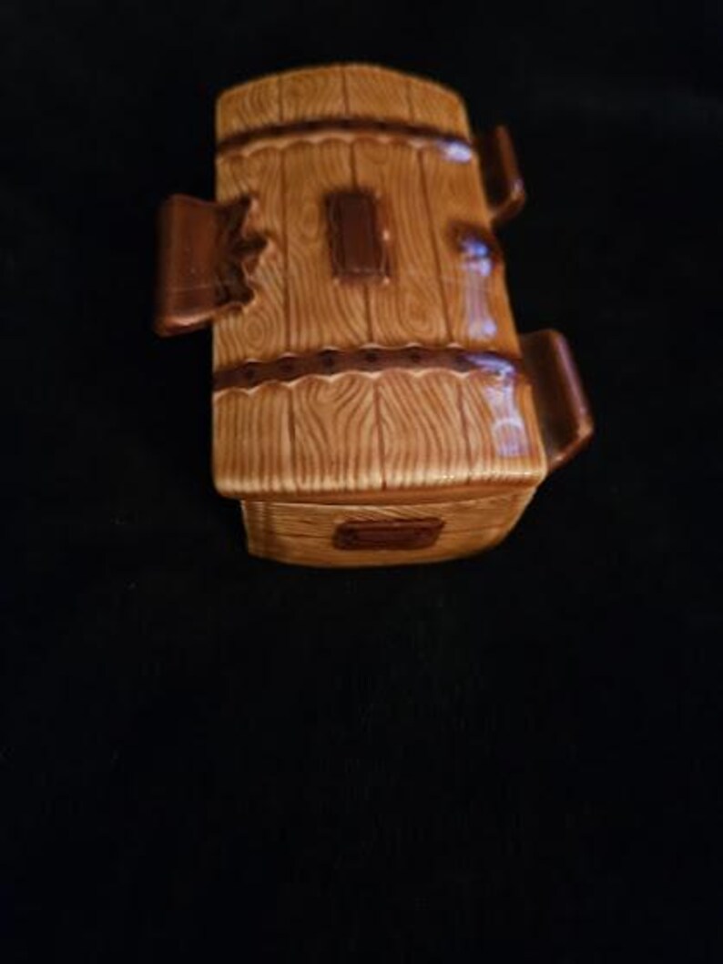 May include: A ceramic barrel-shaped container with a wood-grain design in shades of brown. The container has a rectangular opening and two handles. The design includes dark brown accents and a decorative band around the middle.