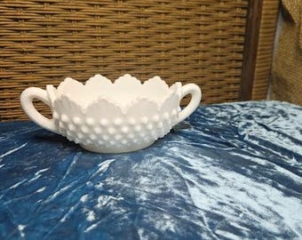 May include: A white hobnail milk glass bowl with two handles. The bowl has a scalloped rim and a textured surface of small, raised bumps. The bowl is on a blue velvet surface, with a woven background.