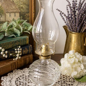 May include: A clear glass oil lamp featuring a rounded, spiraled base and a tall, slender chimney. The lamp includes a metallic gold-toned burner assembly connecting the base to the glass chimney, creating a functional lighting piece.