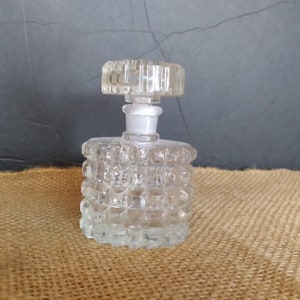 May include: A clear glass bottle with a textured, diamond-shaped design. The bottle has a clear glass stopper with a square top.