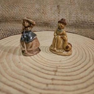 May include: Two small ceramic figurines of people in historical clothing. One figure is wearing a brown dress with a blue apron and a hat. The other figure is wearing a long dress and a hat, holding a bag. Both are on a wooden surface.
