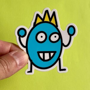 May include: A blue and yellow cartoon character sticker with a crown on its head. The character has a big smile and two large eyes. It is wearing a crown and has two arms raised in the air.