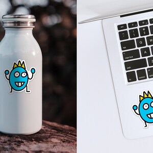 May include: A white stainless steel water bottle with a blue and yellow cartoon sticker of a smiling monster wearing a crown. The monster is raising its arms in the air.