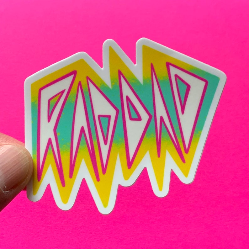May include: A white sticker with the word "RAD" in yellow, pink, and blue letters. The letters are stylized in a jagged, geometric font.