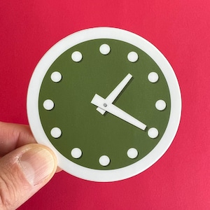 May include: A round clock design with a white border and a dark green face. The clock features white hour markers and hands, indicating the time. The clock is held against a red background.